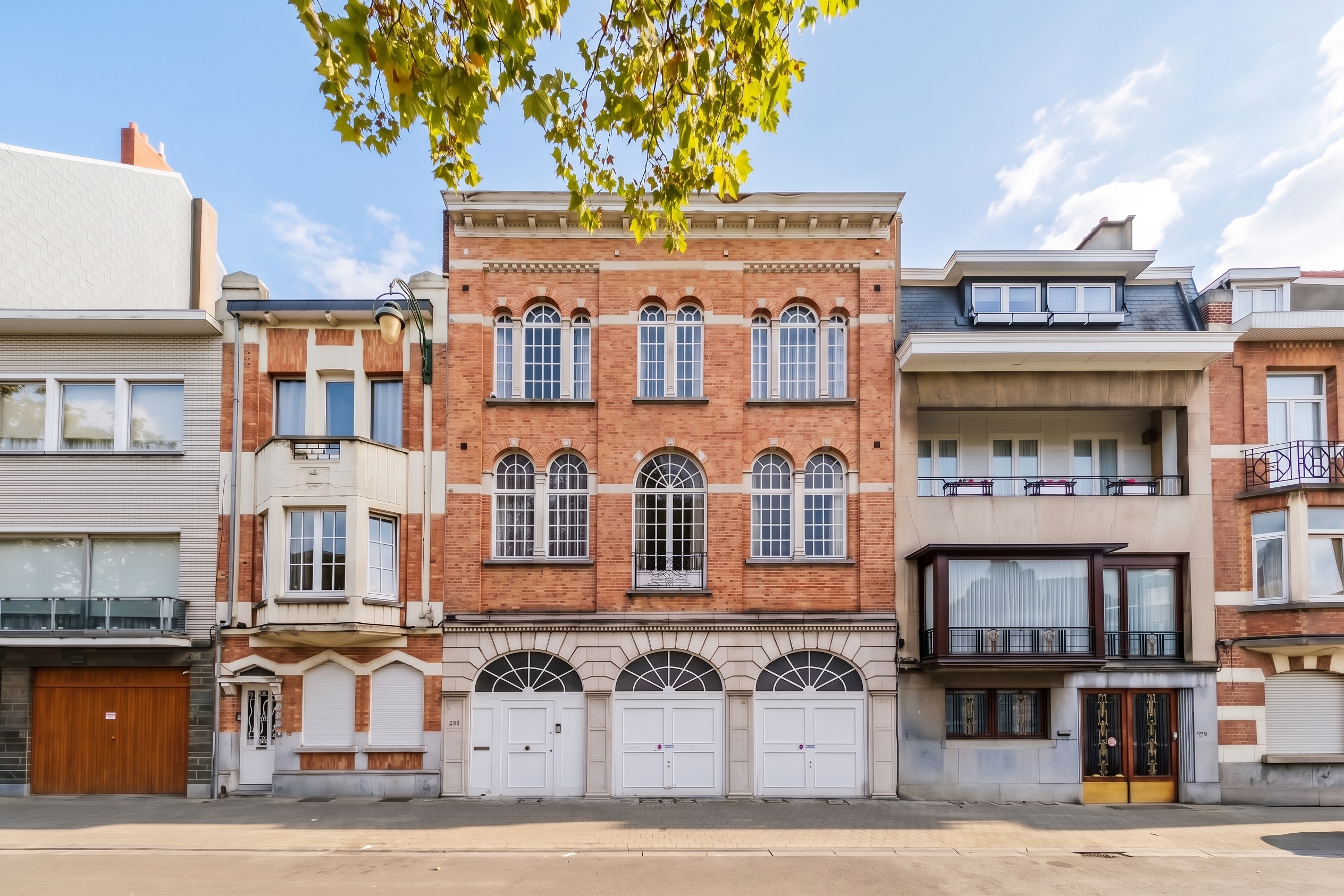 Street view of a historic red brick building with arched windows and white garage doors, flanked by neighboring townhouses. [AI-generated text] [Modifié par IA]