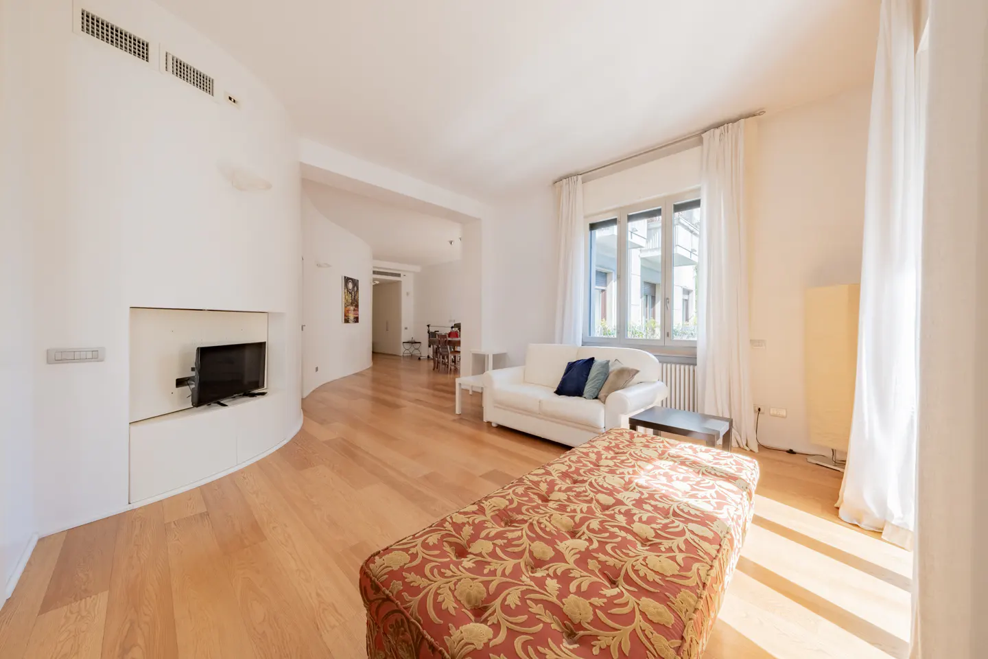 Bright living room with hardwood floors, white walls, and a red patterned ottoman. A white sofa sits near a window with sheer curtains. A TV is mounted in a curved wall.