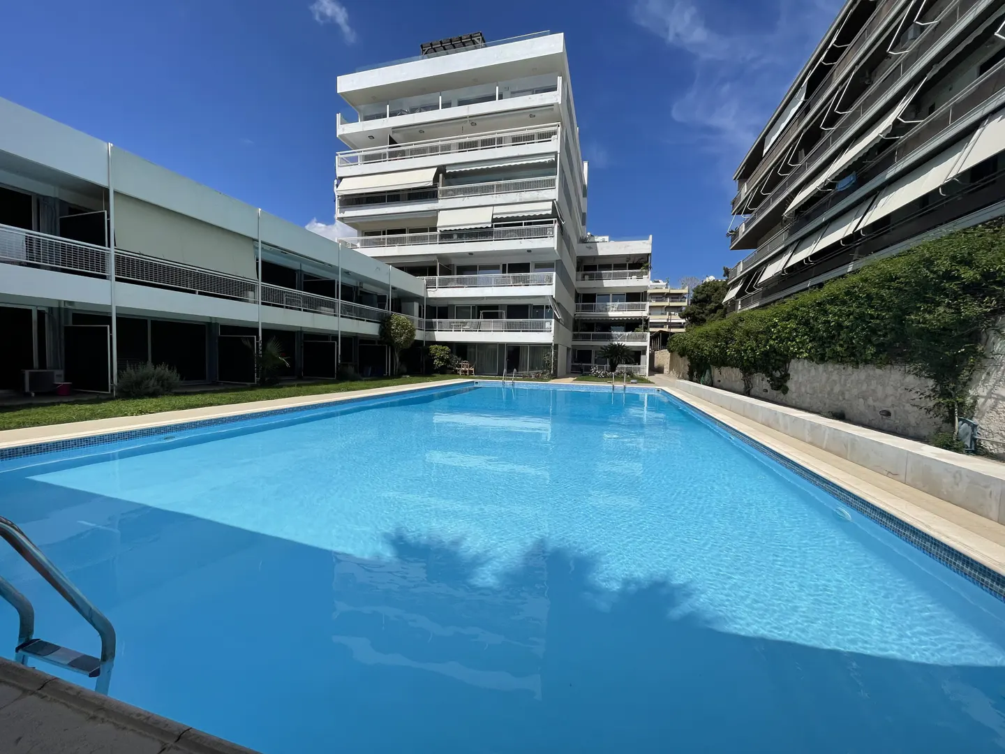 Outdoor pool with clear blue water, surrounded by white apartment buildings under a sunny sky.