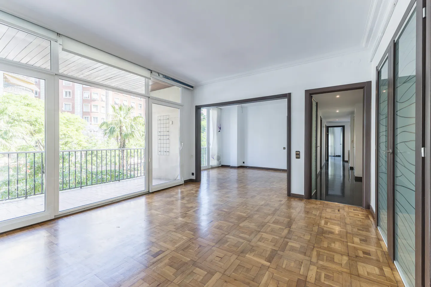 Bright, empty room with parquet floors, white walls, and large windows overlooking a balcony with trees. Dark wood door frames lead to other rooms.