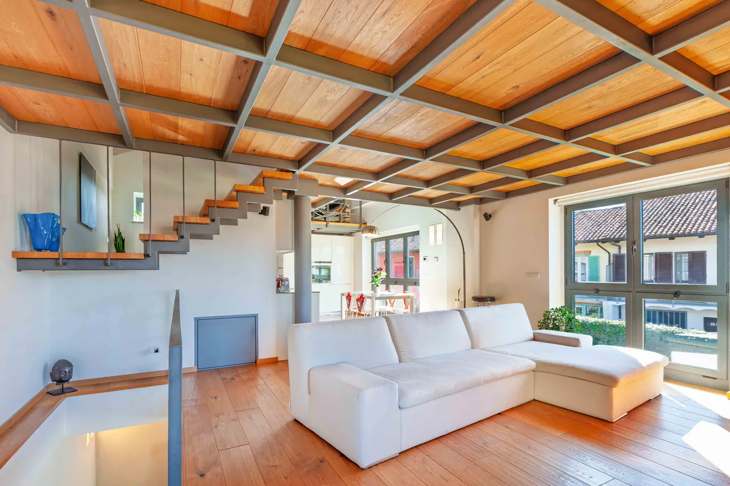 Bright living room with a white sofa, wood floors, and a unique wood and metal grid ceiling. Stairs lead to a loft area. Large windows offer natural light.