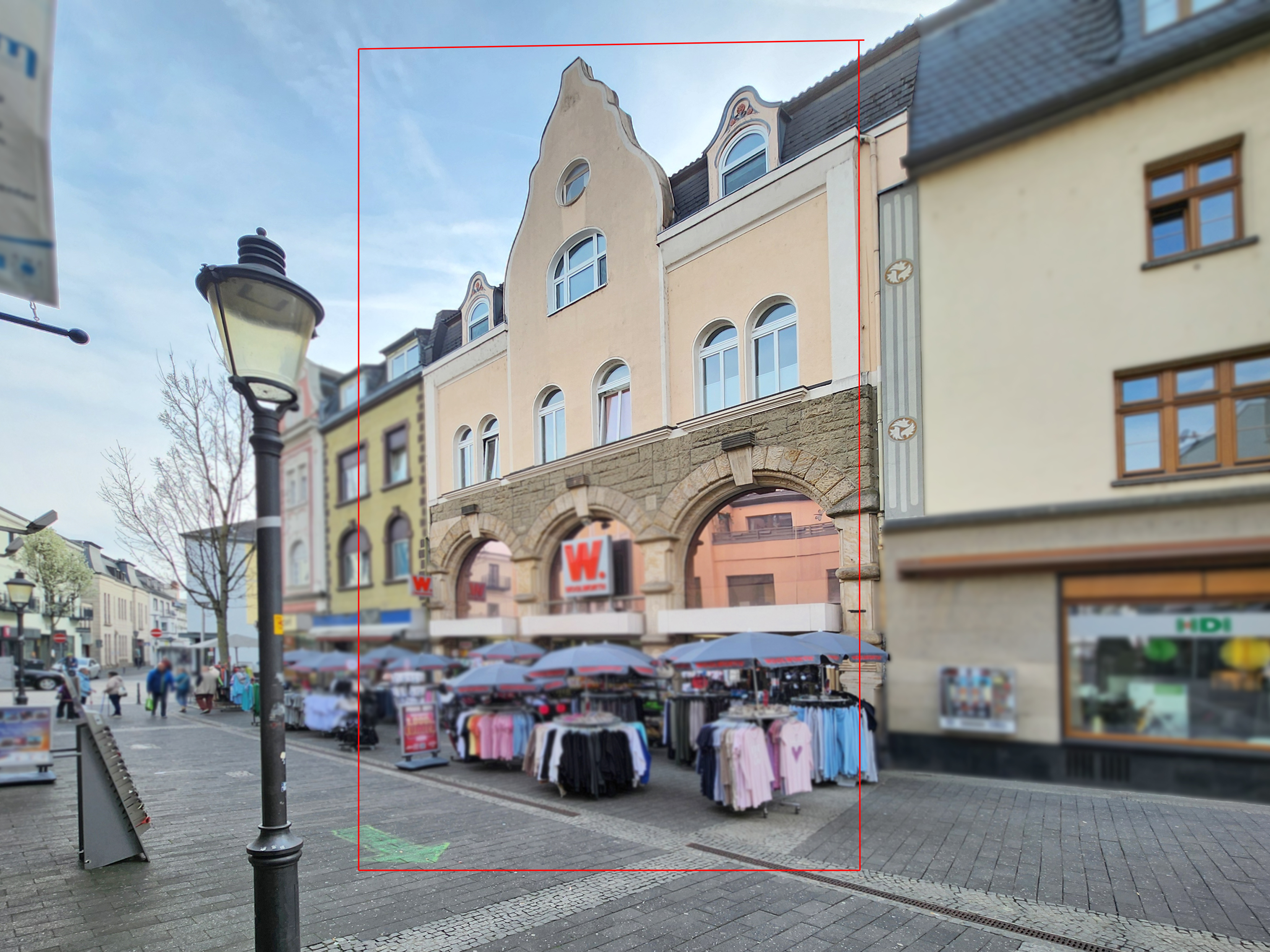 Street view of a three-story beige building with arched openings above market stalls with clothing under blue umbrellas.