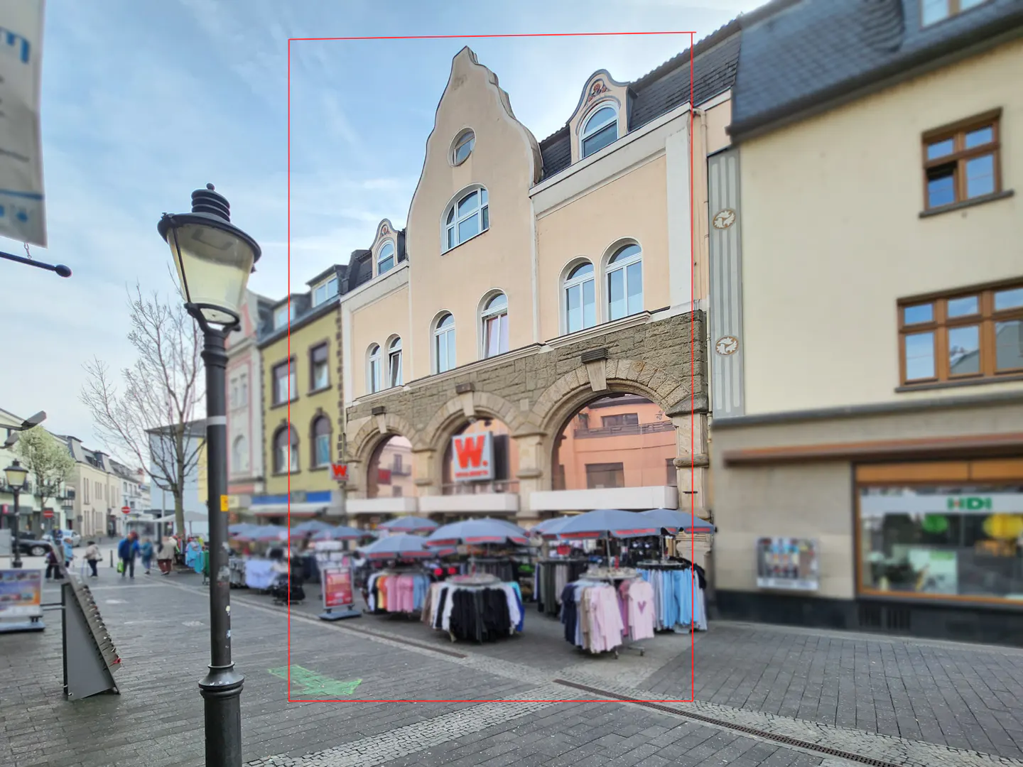 Street view of a three-story beige building with arched openings above market stalls with clothing under blue umbrellas.