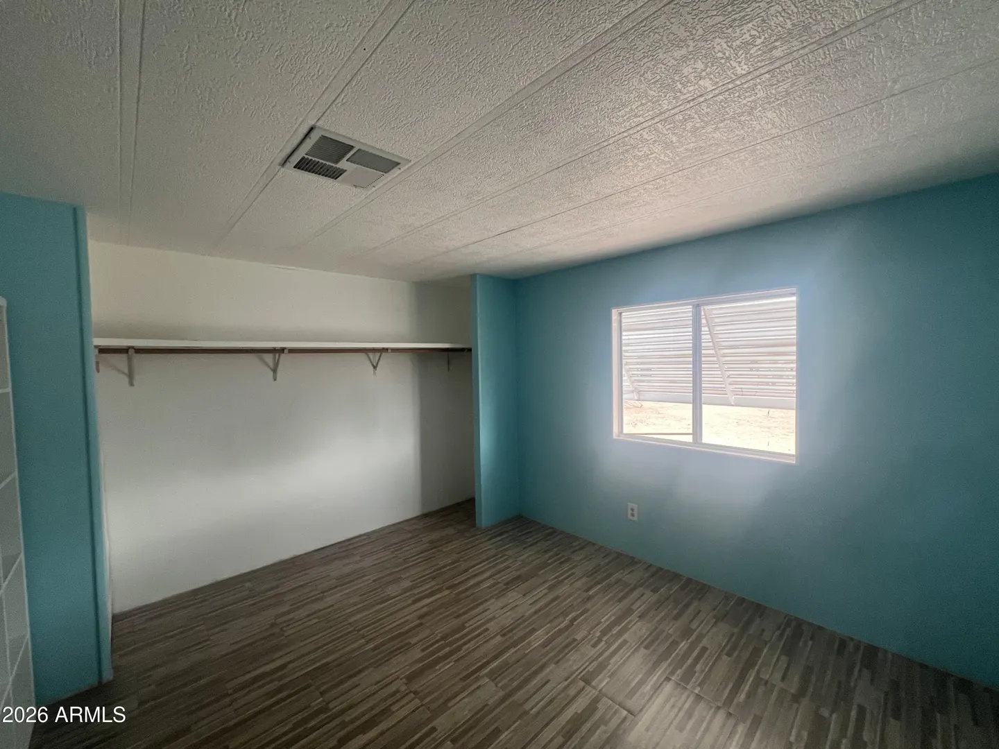 A bedroom with blue walls, wood-look tile flooring, and a white closet with a shelf. A window lets in natural light.
