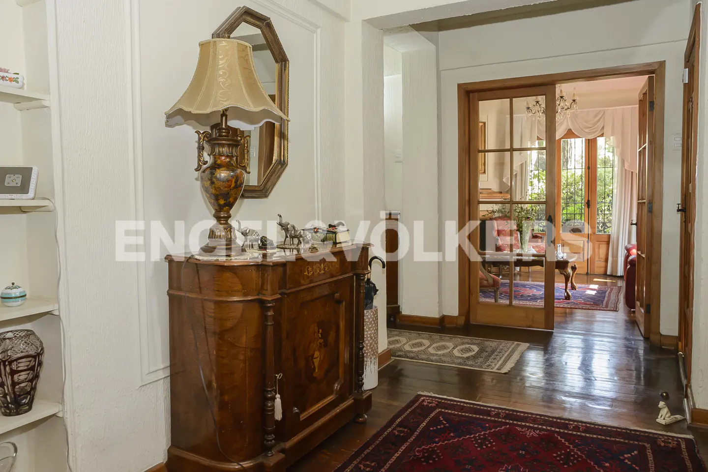 A foyer with a wooden cabinet, lamp, and mirror. A doorway leads to a living room with a chandelier and view of a garden.