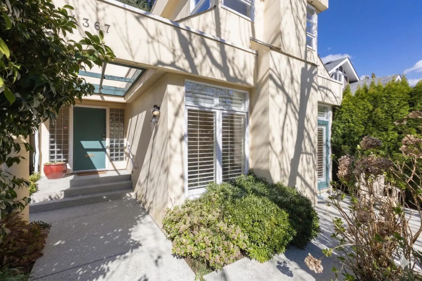 Exterior view of a two-story beige house with a green front door, glass block sidelights, and white shuttered windows.