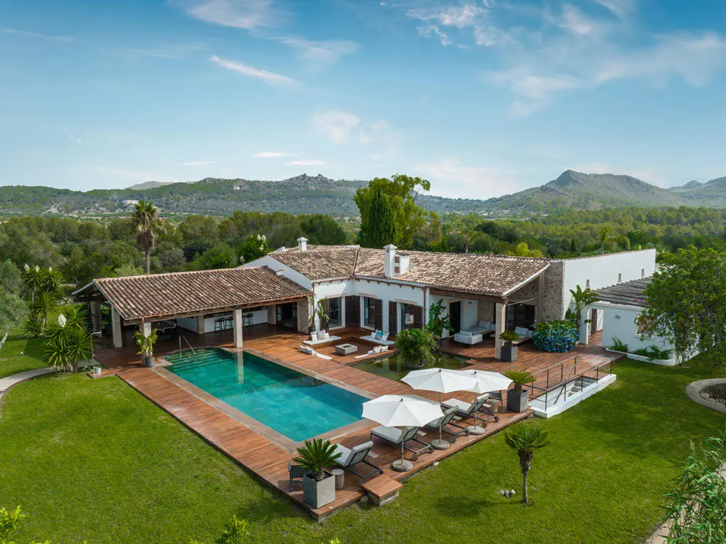 Aerial view of a white villa with a pool, wooden deck, lounge chairs, and umbrellas, surrounded by green lawn and trees. Mountains in the background.