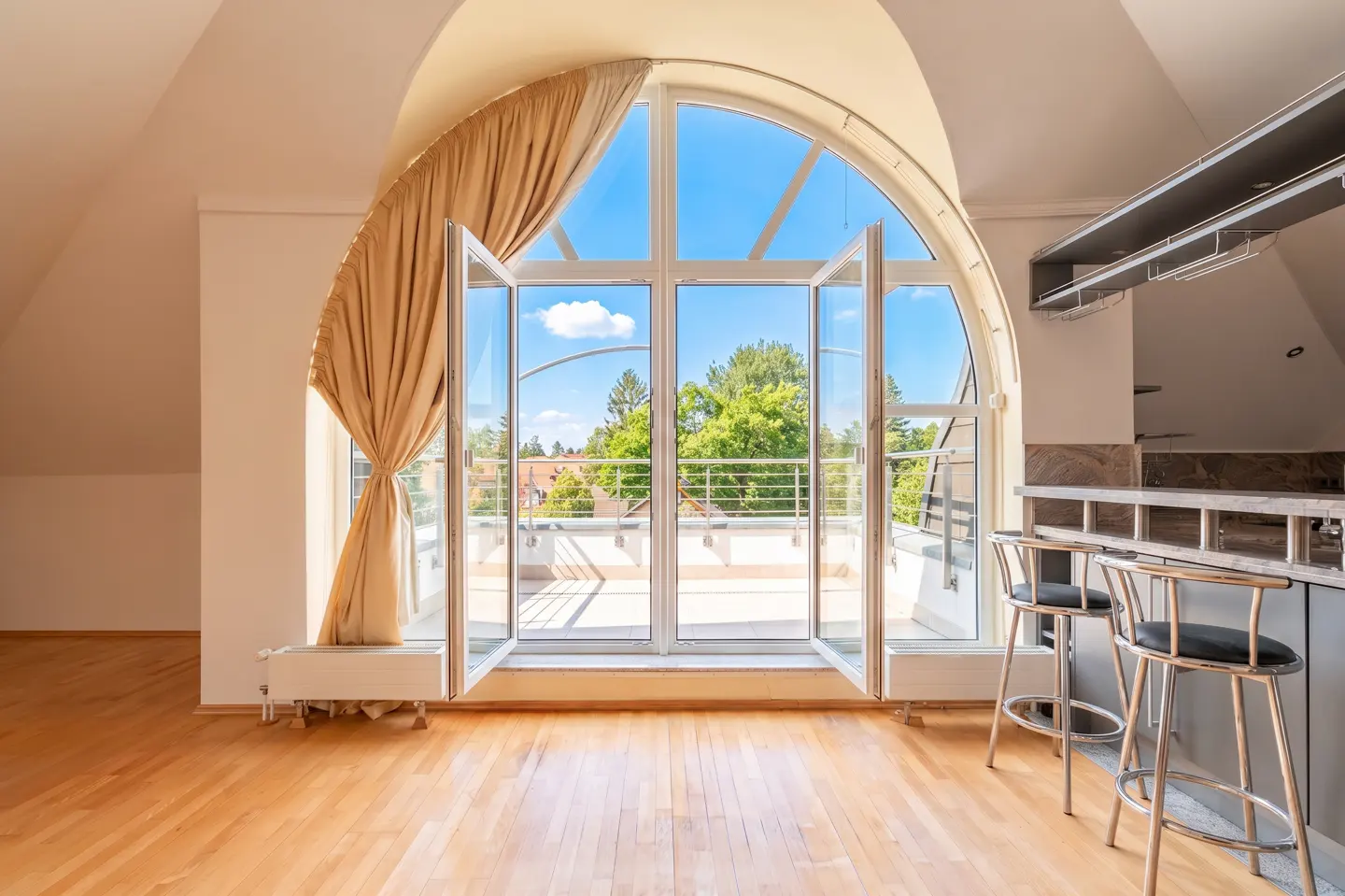 Bright room with wood floors, arched window open to a balcony, and a bar with two stools. A tan curtain hangs on the left side of the window.