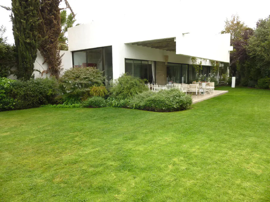 Modern white house with large windows, patio furniture, and a green lawn. Lush greenery surrounds the property.