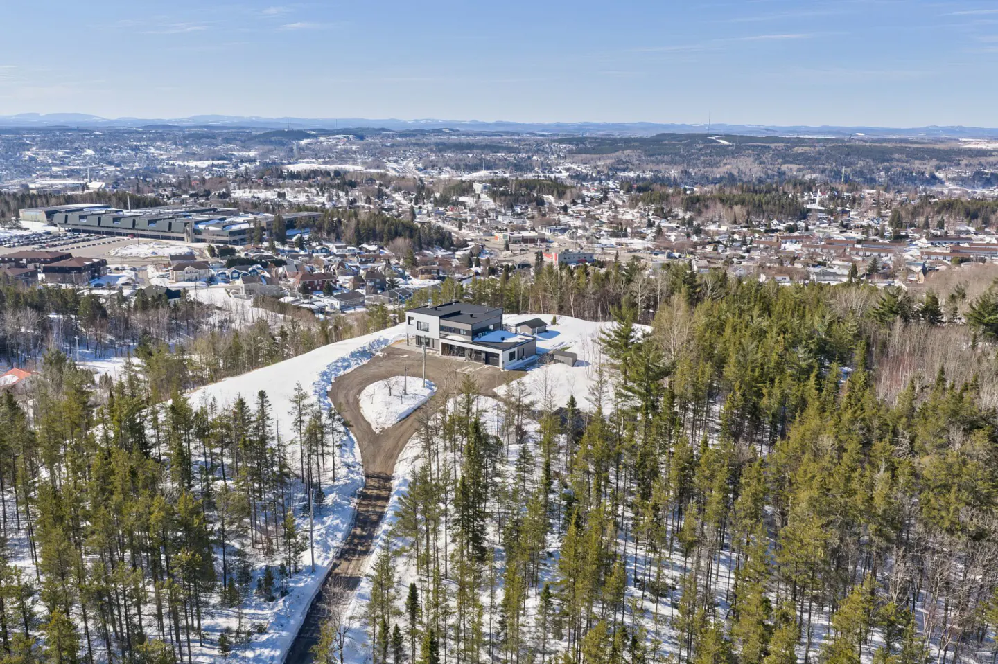 Aerial view of a modern black and white house on a snow-covered hill, surrounded by green trees, with a town in the background.