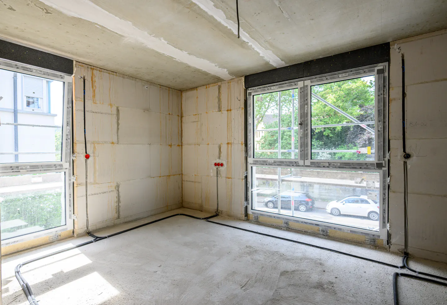 Unfinished room with concrete walls, floor, and ceiling. Black wires run along the baseboards and walls. Two large windows show a street scene.