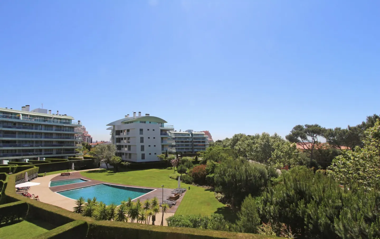 View of modern white apartment buildings with a pool, green lawn, and trees under a clear blue sky.