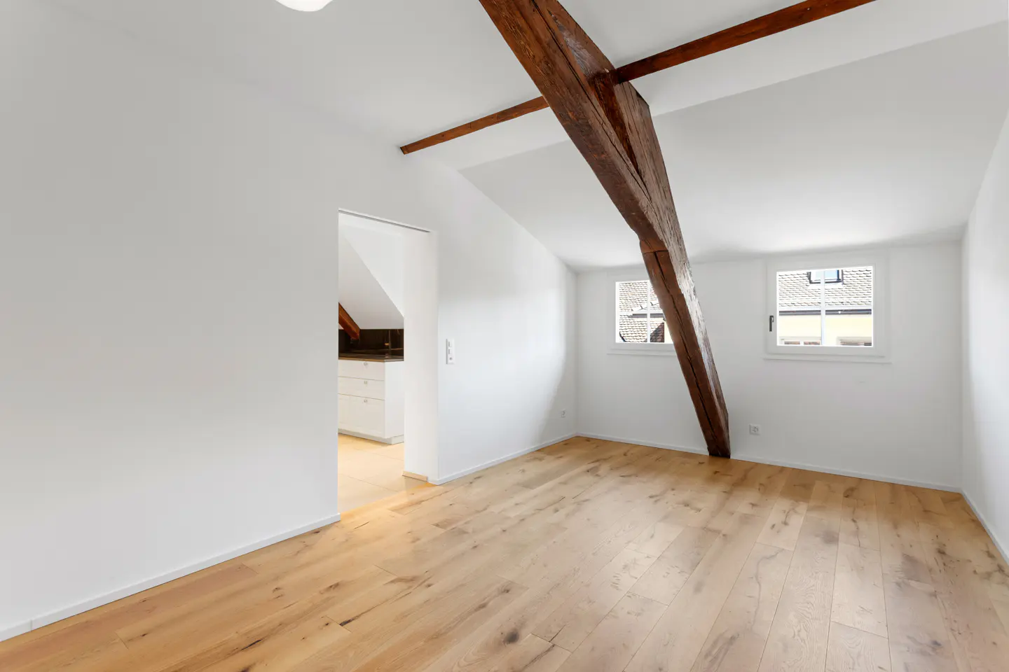 Bright, empty room with wood floors, white walls, and exposed beams. Two windows let in natural light. A doorway leads to another room.