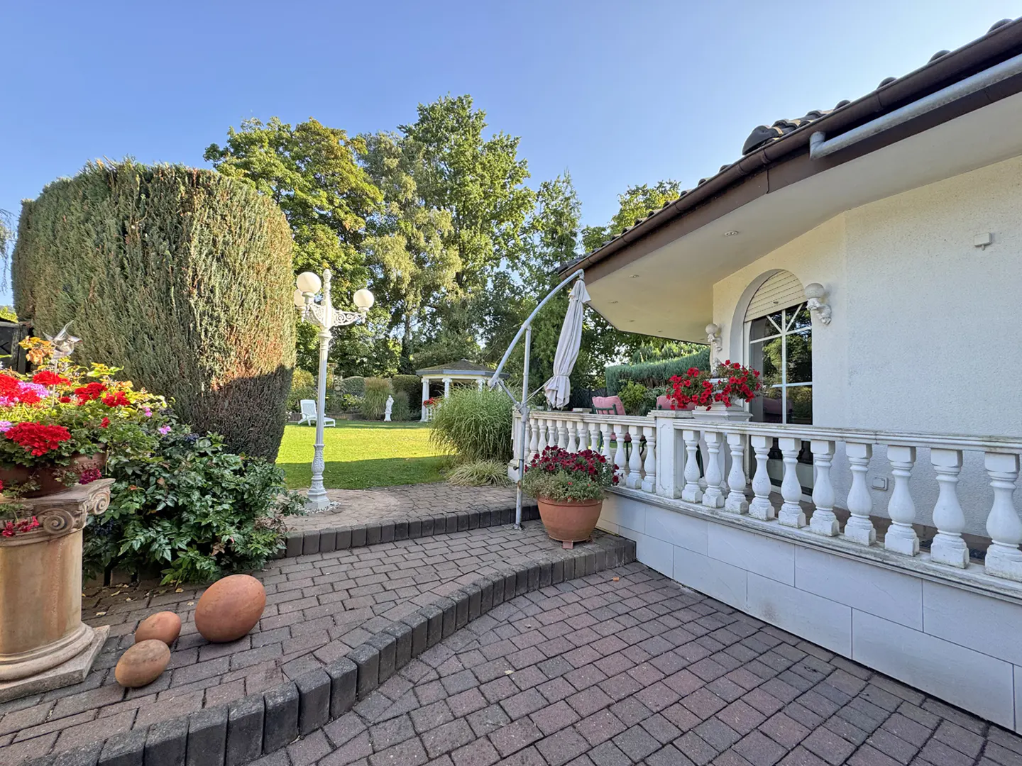 Brick patio leads to a white house with a balustrade. Red flowers decorate the patio. A green lawn and trees are in the background.