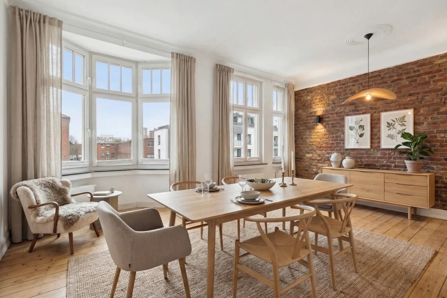 Bright dining room with wood floors, a brick accent wall, and a large bay window with beige curtains. A light wood table is centered on a jute rug.