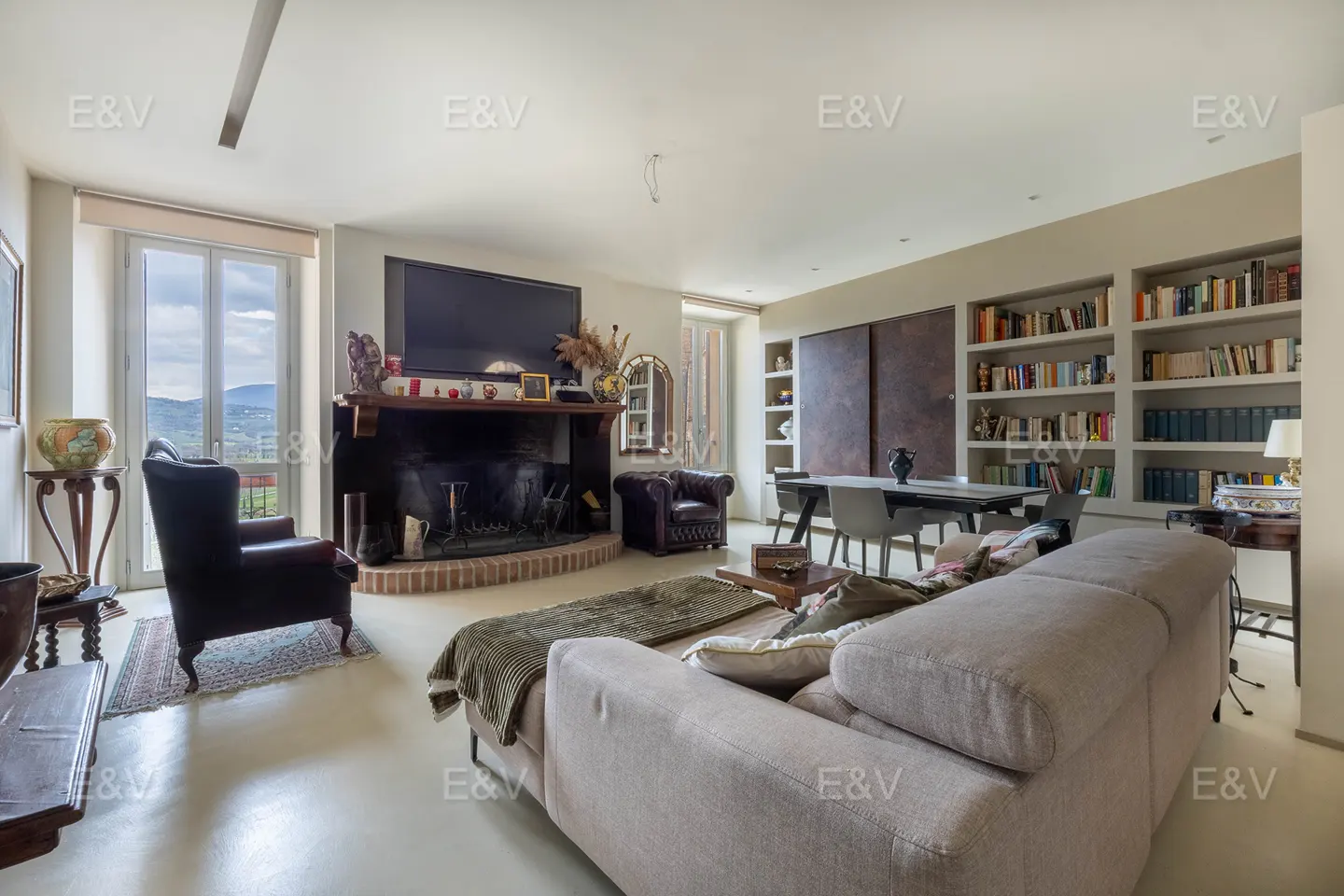 Living room with a beige sofa, fireplace, TV, bookshelves, and a dining table with chairs. A window shows a view of the hills.