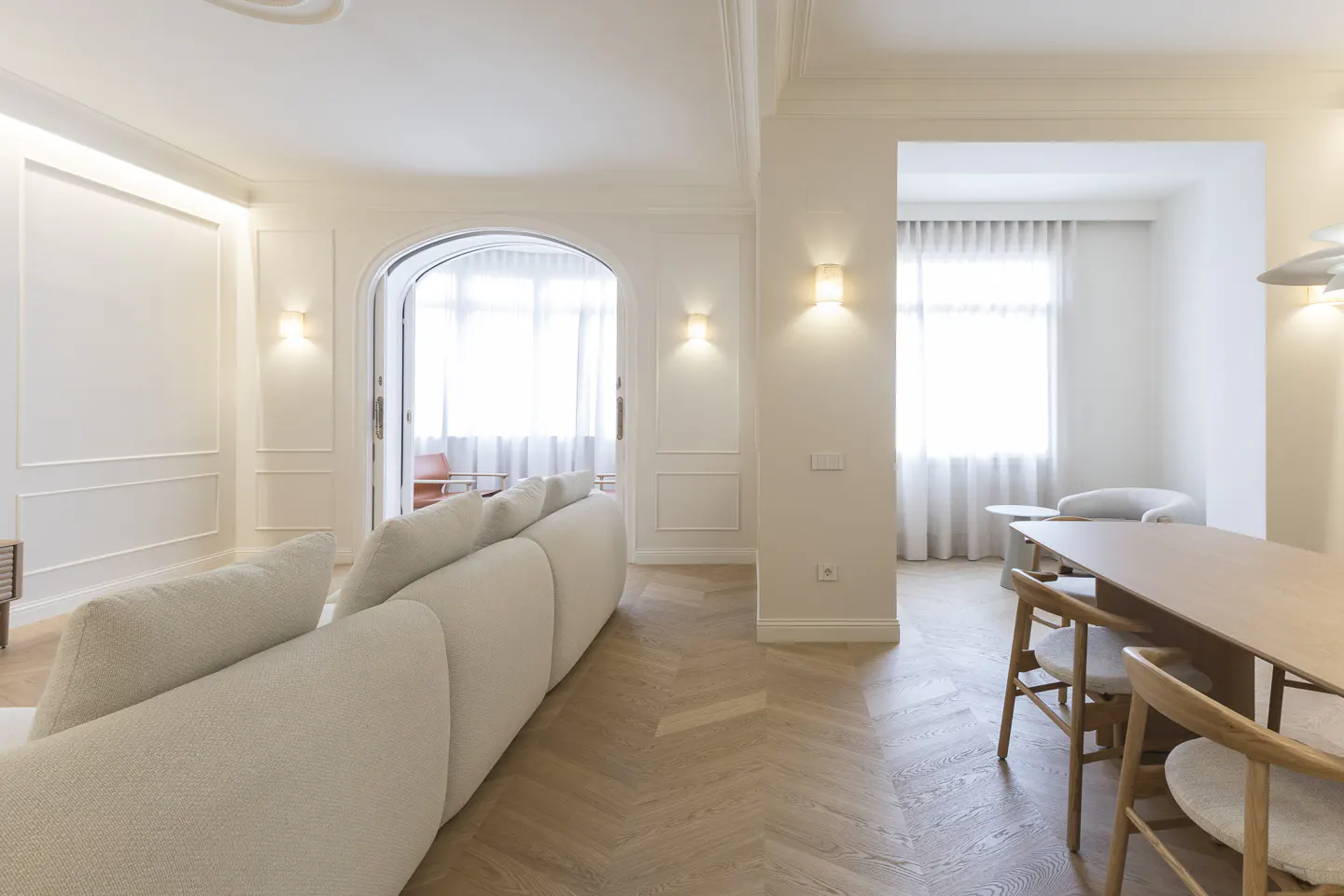 Bright, modern living room with a white sofa, wood floors, and a dining table with chairs. Arched doorway leads to a balcony.