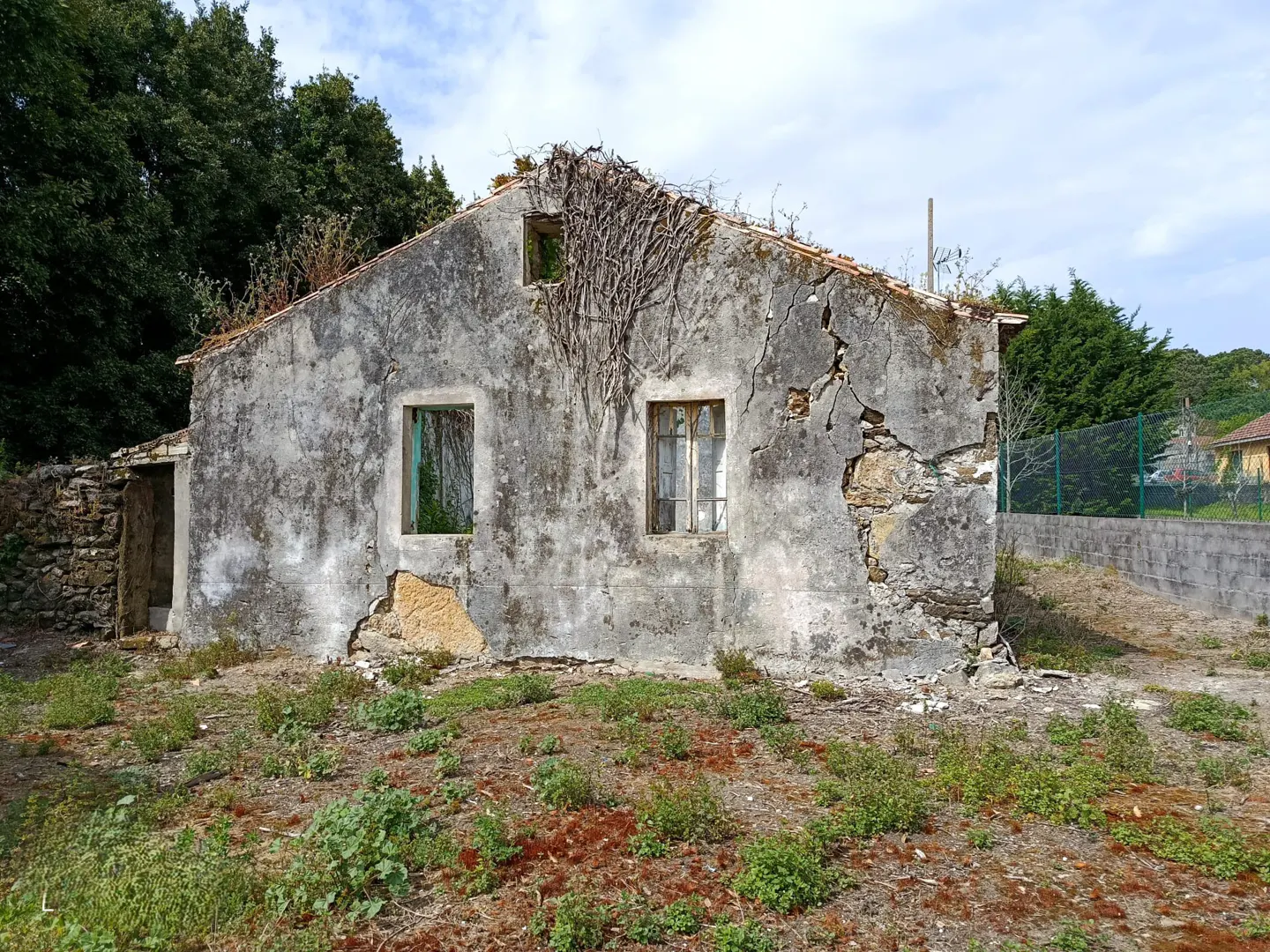 Exterior of a dilapidated, one-story house with a cracked, gray facade and overgrown vegetation. Two windows and a doorway are visible.