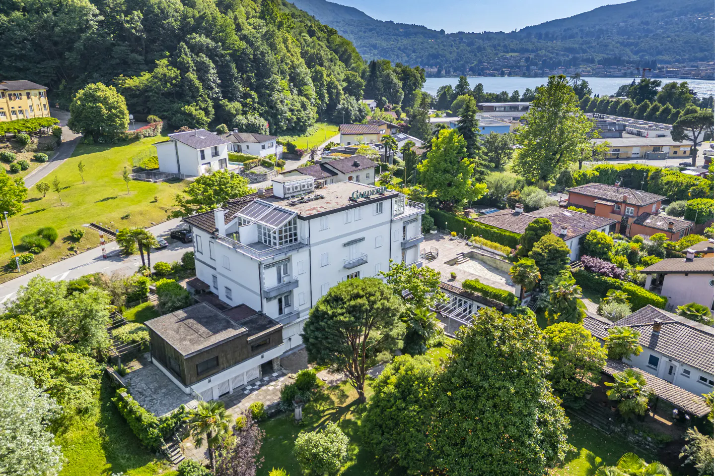 Aerial view of a white multi-story building surrounded by lush green trees, gardens, and nearby homes near a lake.