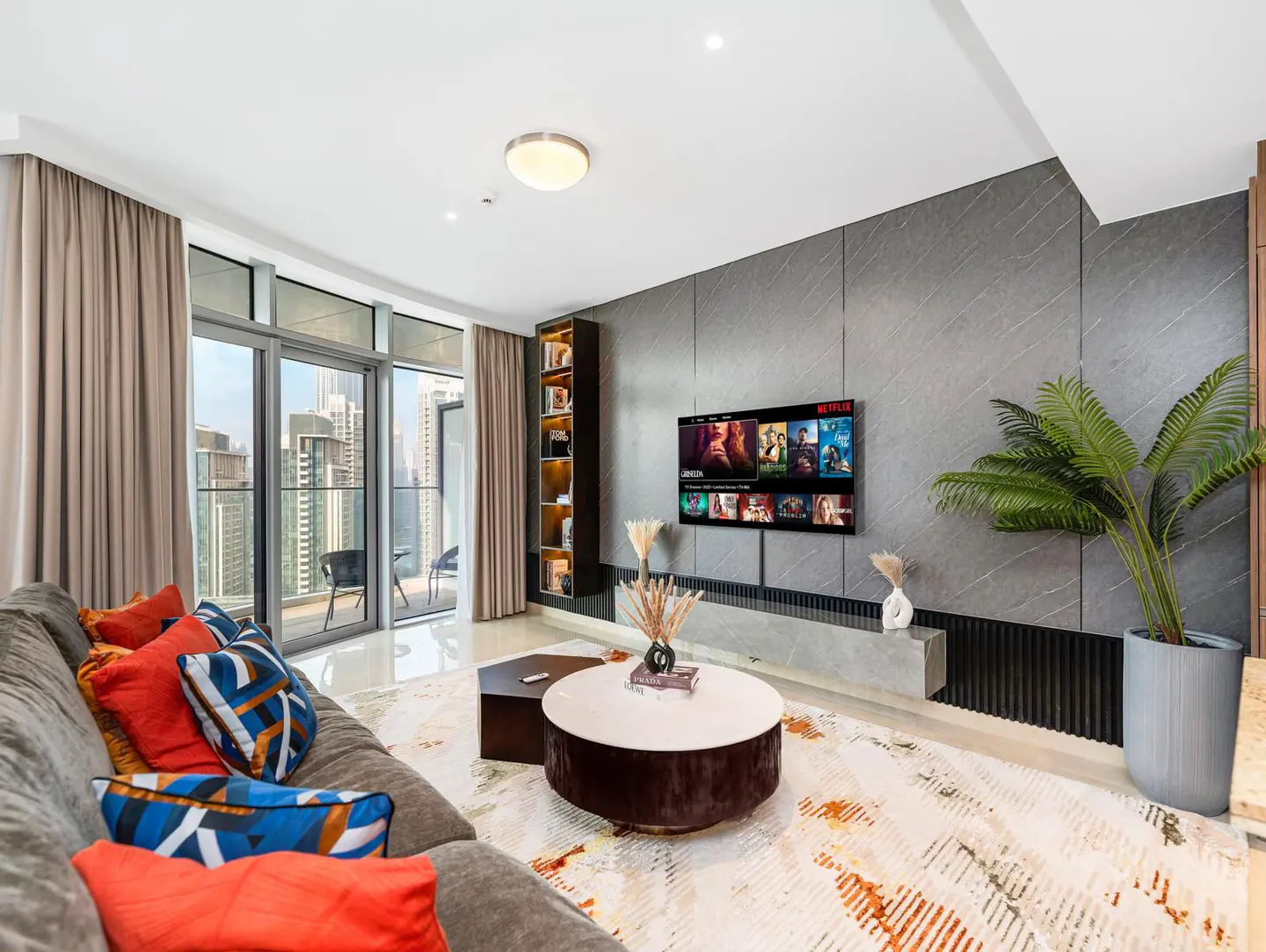 Living room with a gray sofa, colorful pillows, a round coffee table, and a TV showing Netflix. A balcony overlooks a city skyline.