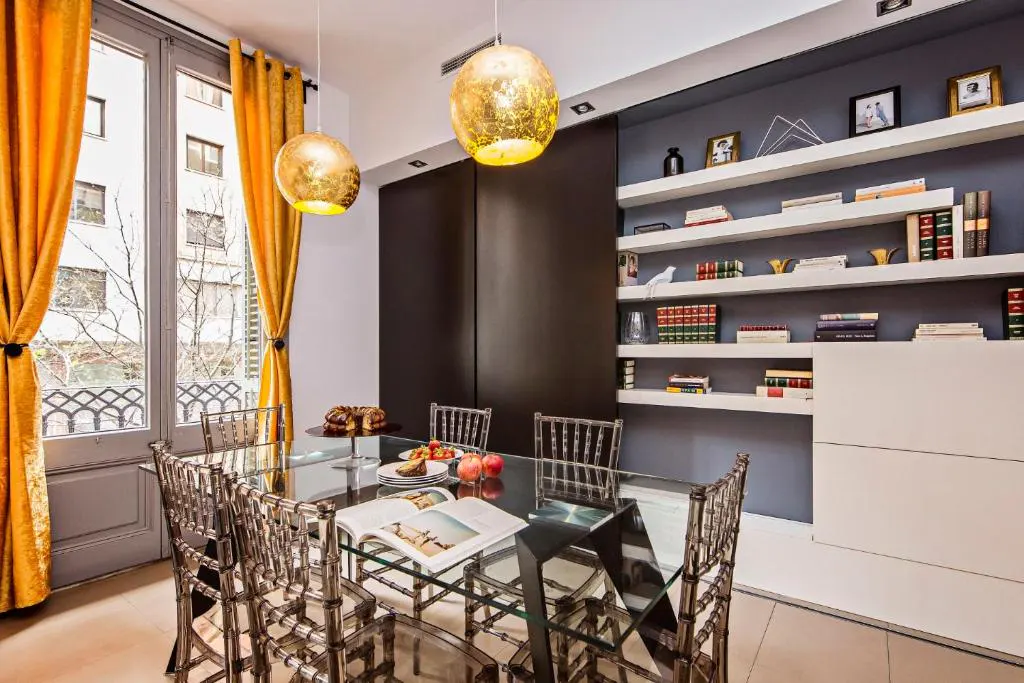 Dining room with glass table, clear chairs, and gold pendant lights. Shelves with books and decor are on the back wall. Gold curtains frame the window.