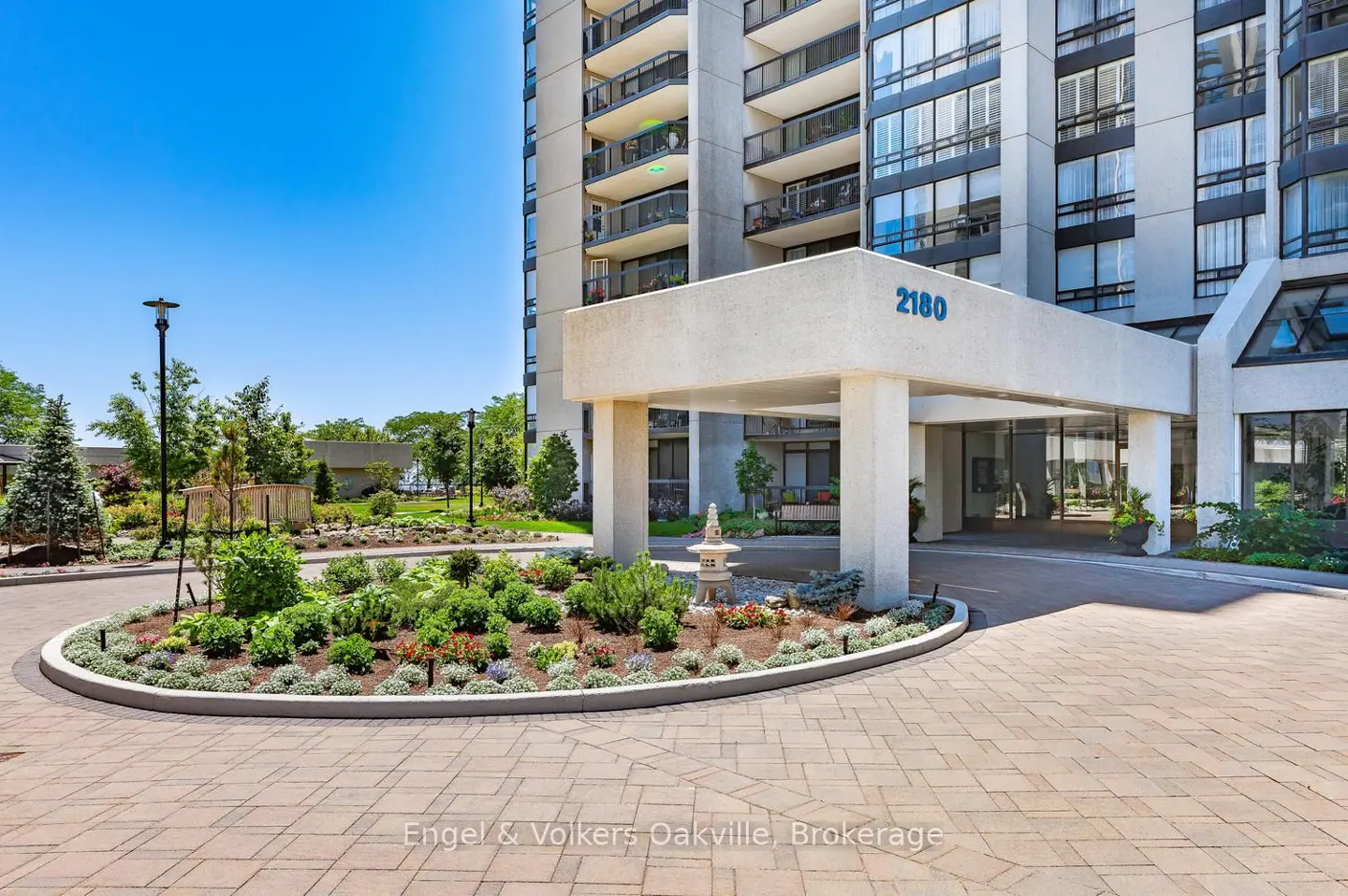 Exterior view of a high-rise condo building with a covered entrance, landscaping, and a brick driveway on a sunny day.