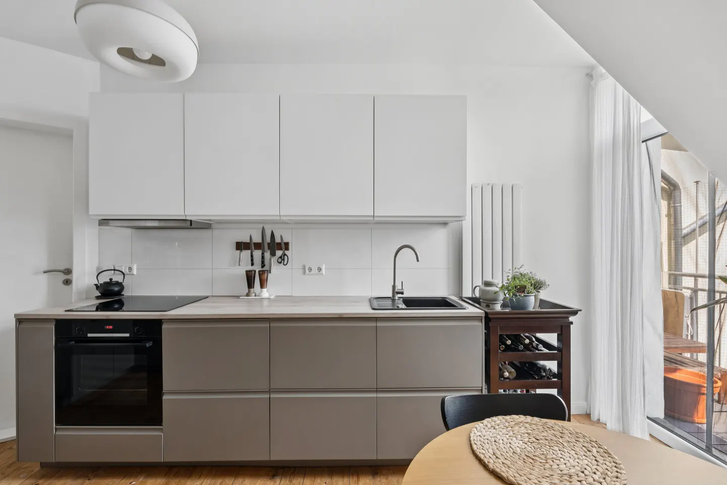 Modern kitchen with white upper cabinets, taupe lower cabinets, black sink, and a round wooden table with a woven placemat. Balcony in background.