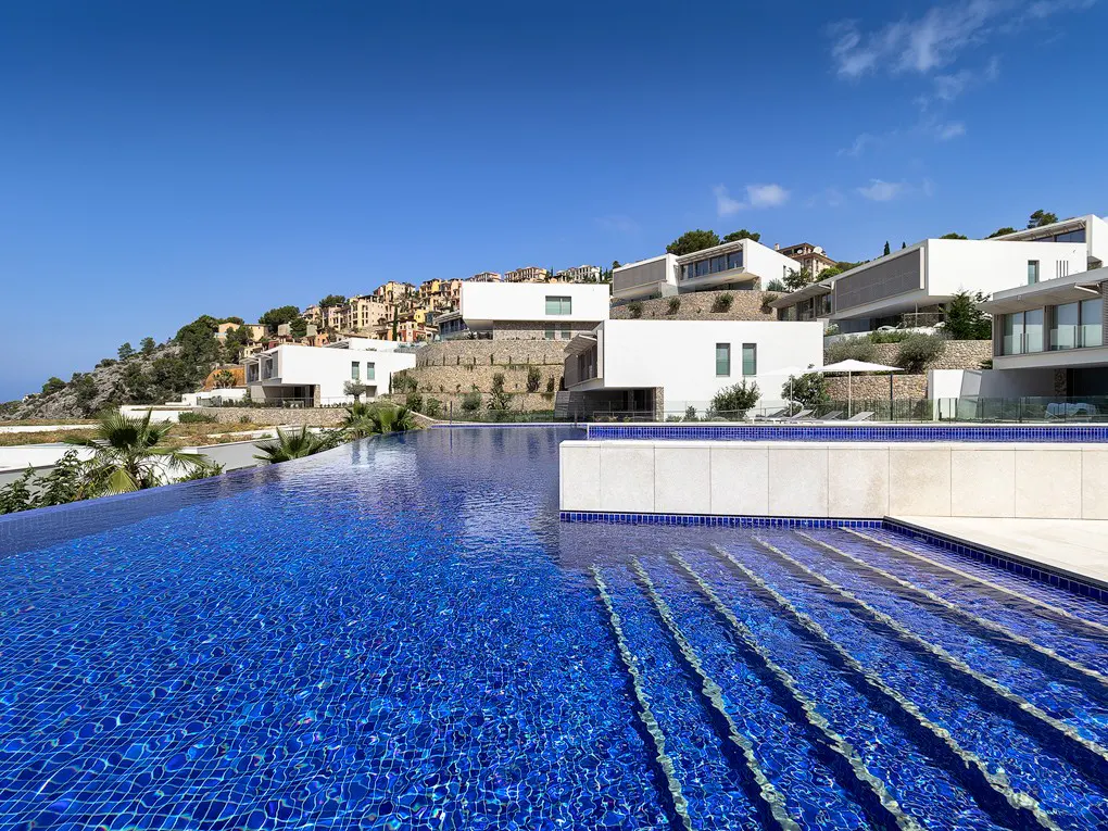 A luxurious infinity pool with blue tiles leads to modern white villas on a hillside under a clear blue sky.