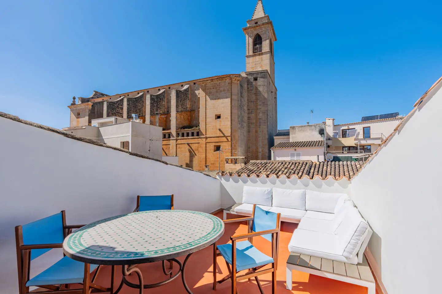 Rooftop patio with blue chairs, table, and white sofa. A stone church with a bell tower is in the background under a clear blue sky.