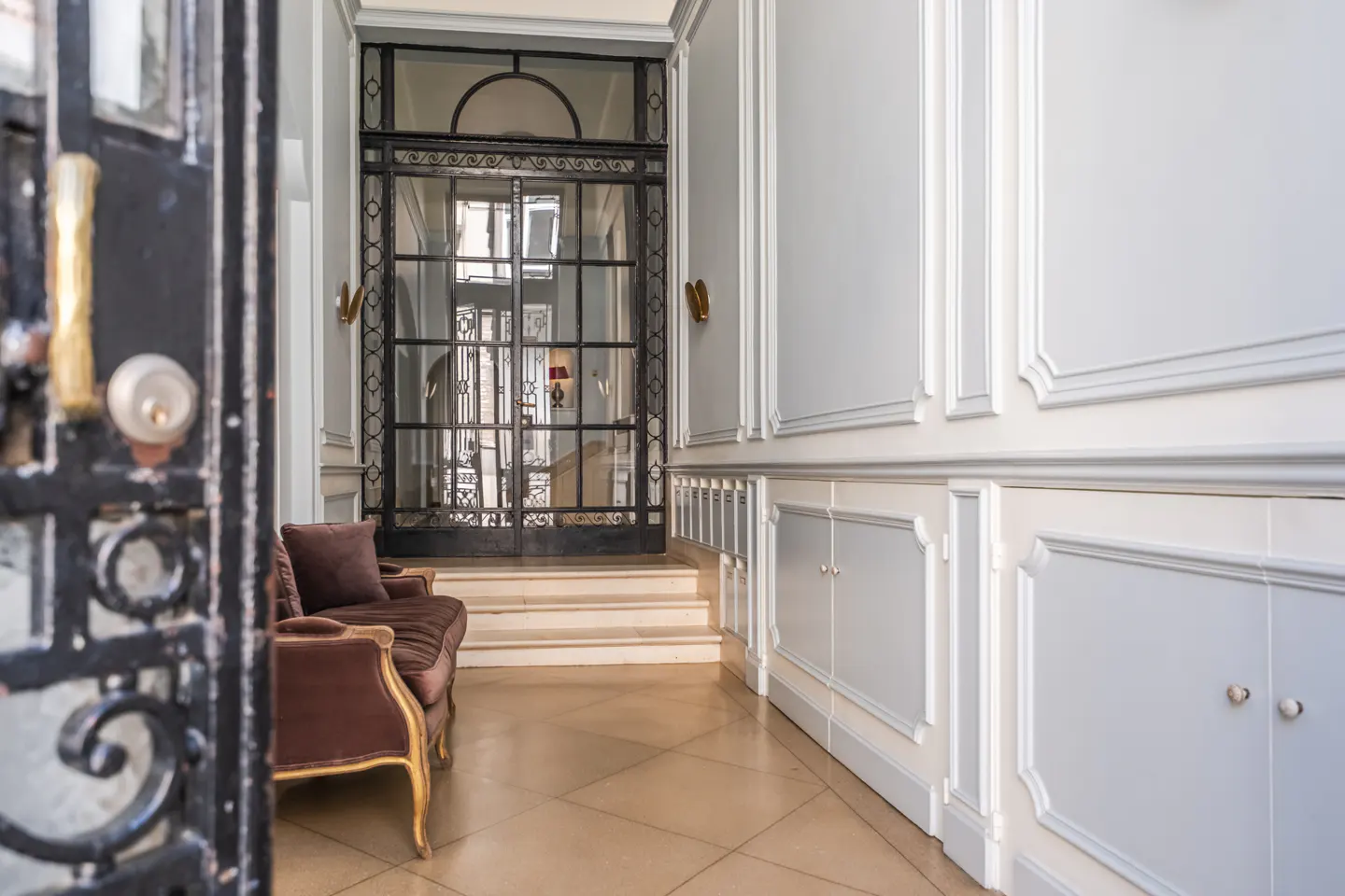 Elegant foyer with a brown velvet sofa, black iron gate, and light blue paneled walls. A tiled floor leads to a glass door with stairs beyond.
