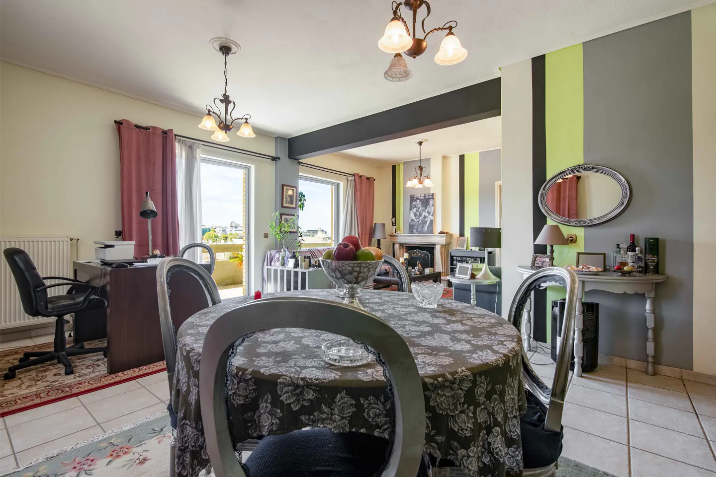 A bright dining room with a round table covered in a floral tablecloth, silver chairs, and a view of the city.