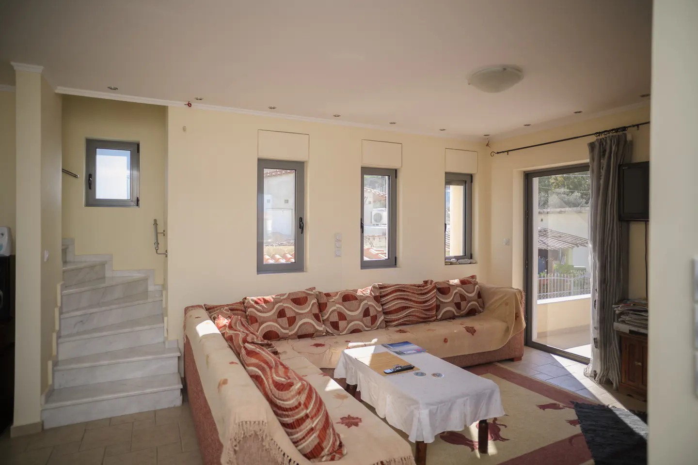 Living room with beige walls, a large sectional sofa with patterned pillows, and a white coffee table. Stairs and windows are visible.