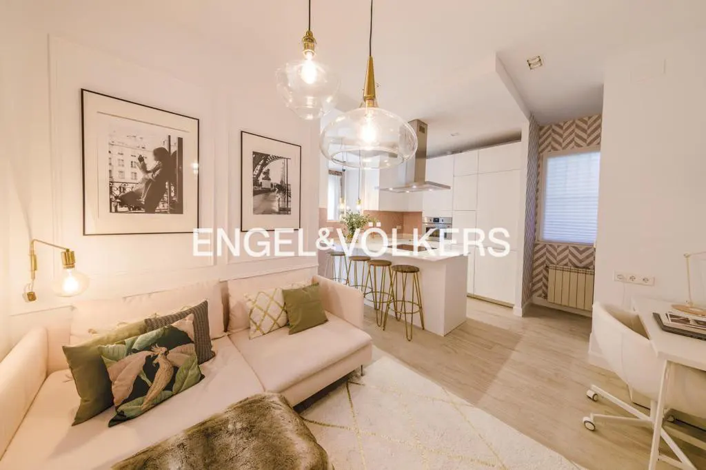 A bright, modern apartment interior featuring a pink sofa with decorative pillows, a kitchen island with stools, and black and white framed art.