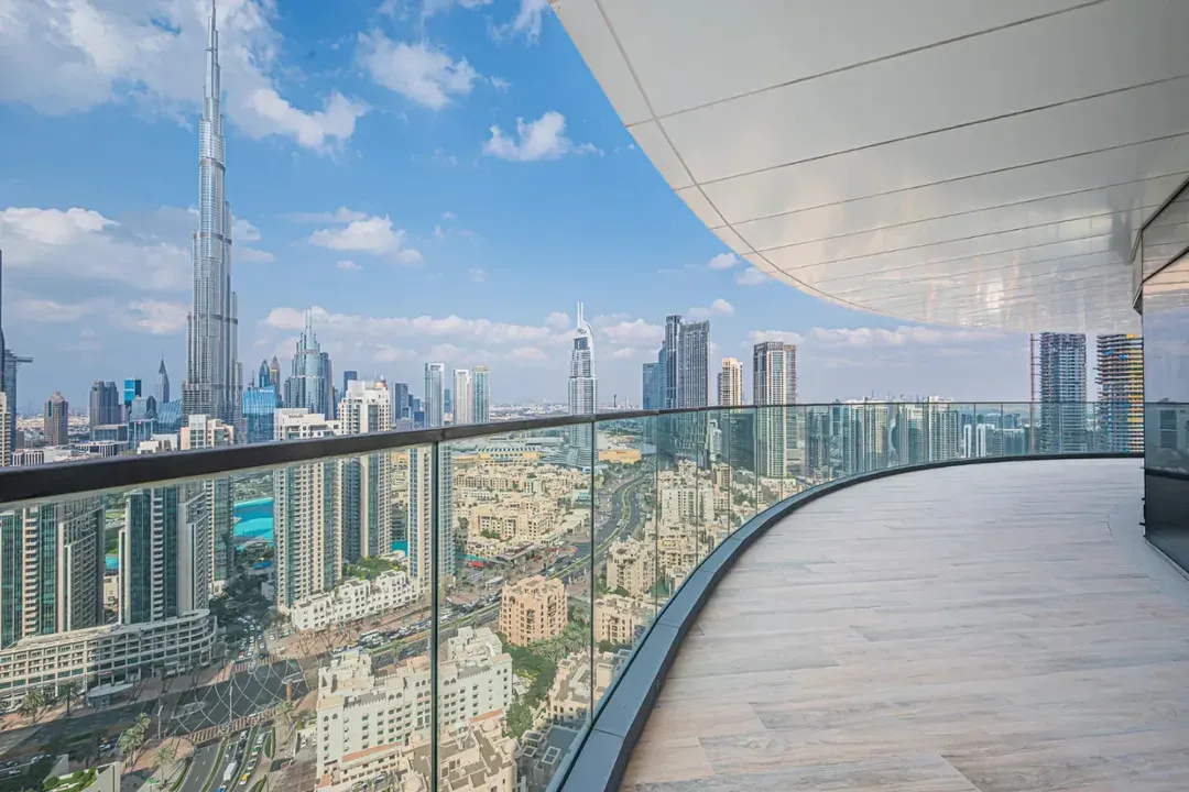View from a curved balcony with glass railings overlooking the Dubai skyline, including the Burj Khalifa, on a sunny day.