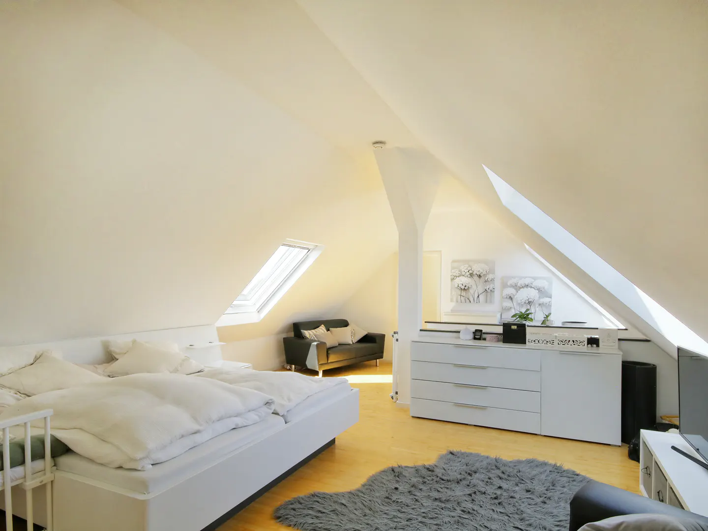 Bright attic bedroom with white walls, a skylight, and a white bed. A gray rug sits on the wooden floor. A black sofa and white dresser are also visible.
