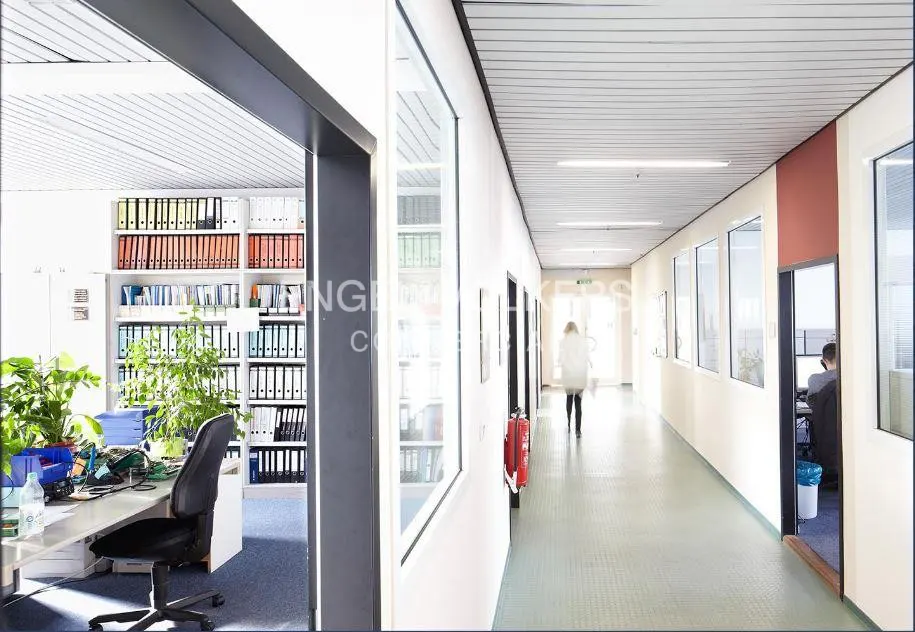 Office interior with a long hallway, white walls, and a woman walking away. An office with shelves and plants is visible on the left.
