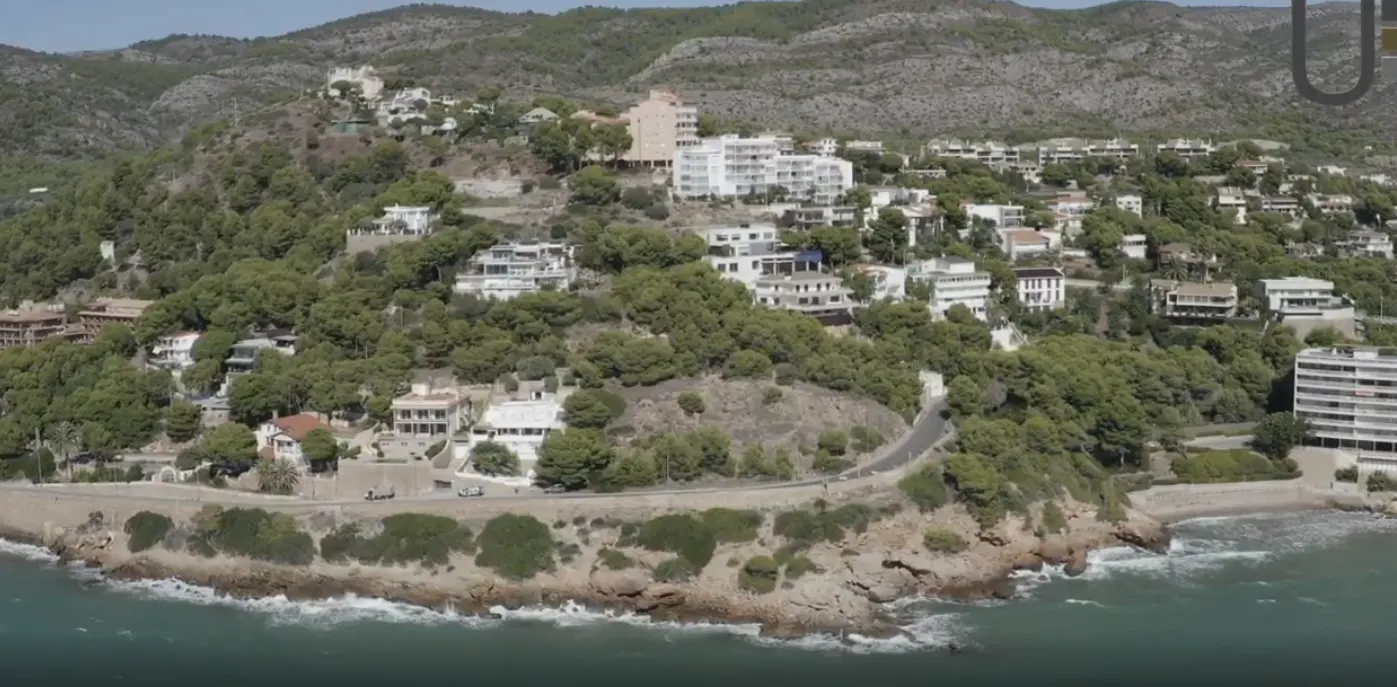 Coastal hillside with white houses and green trees overlooking the ocean. A road winds along the rocky shoreline.