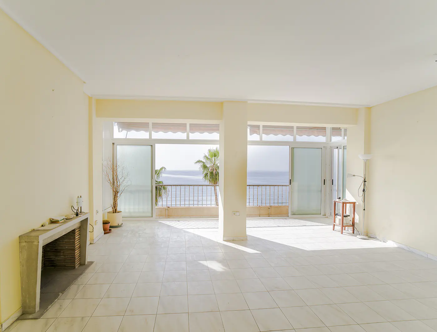 Bright, empty room with cream walls and tiled floor. Sliding glass doors open to a balcony with ocean view and palm trees. Fireplace on the left.