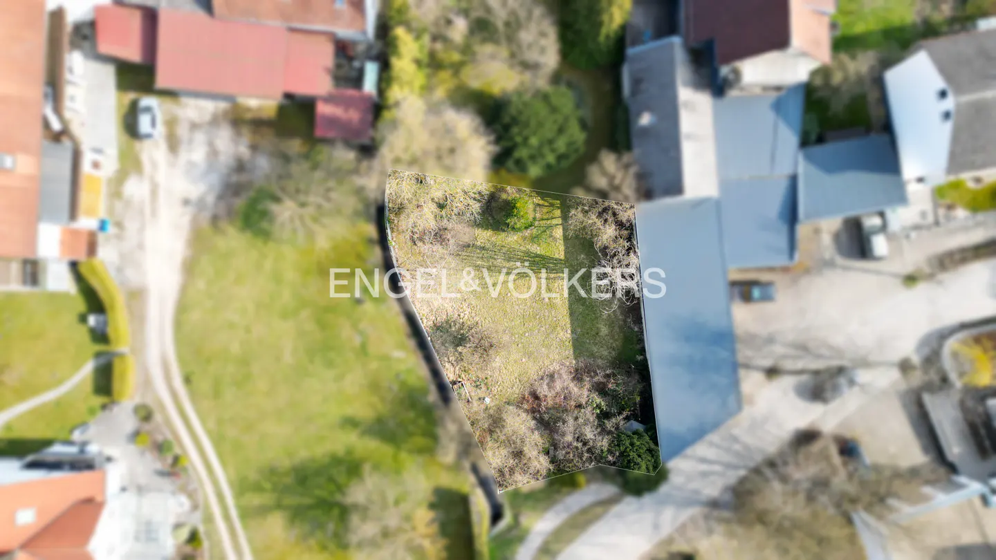 Aerial view of a vacant lot with green grass and some trees, surrounded by buildings and roads. Engel & Völkers logo is superimposed.
