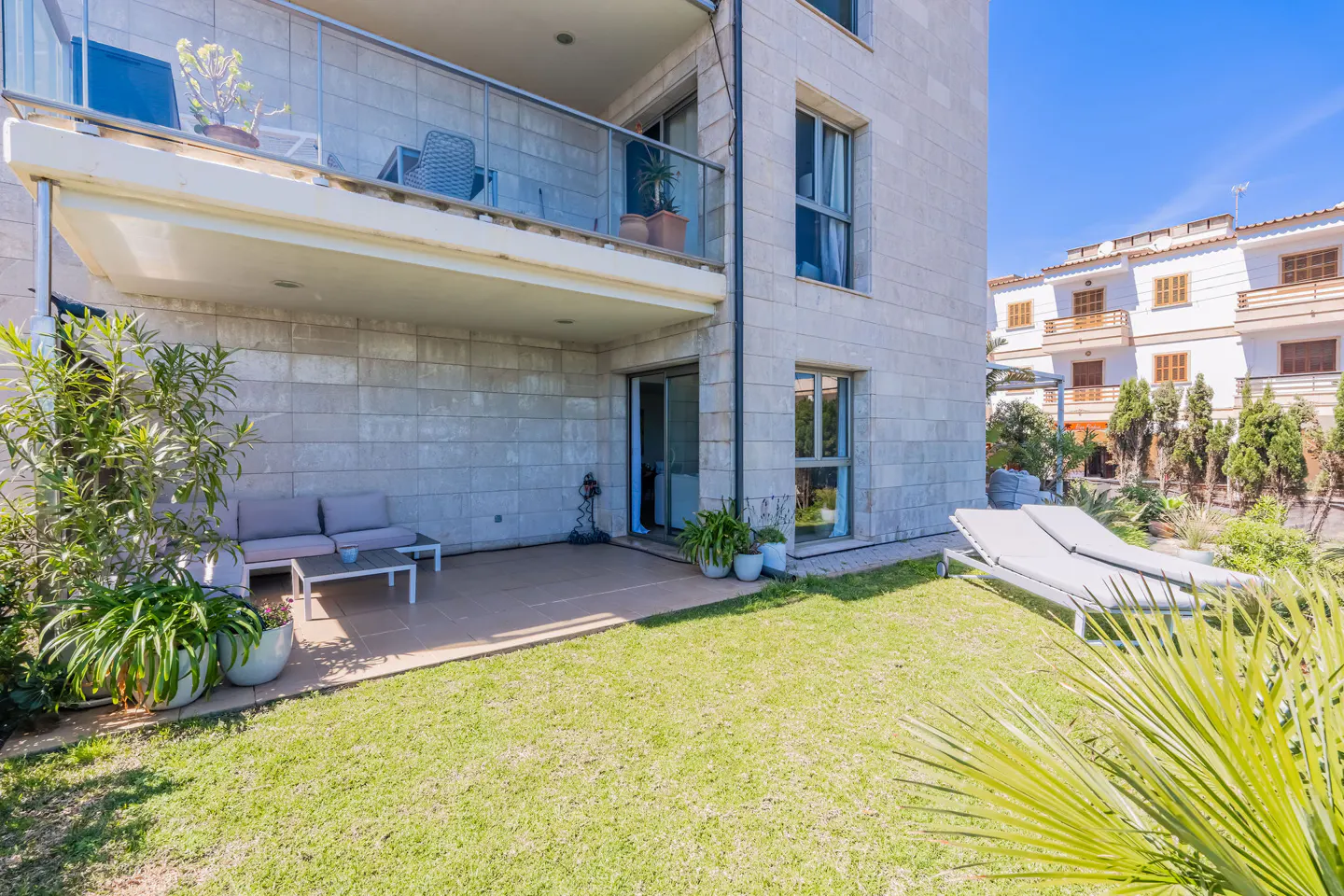Exterior view of a stone building with a balcony, patio, and lawn. Lounge chairs sit on the grass, with plants and other buildings in the background.