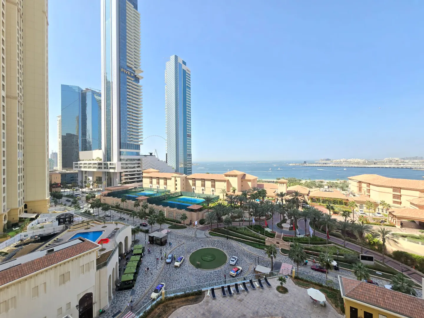 View of Dubai Marina with skyscrapers, hotels, and the Arabian Gulf under a clear blue sky. Palm trees and a roundabout are in the foreground.