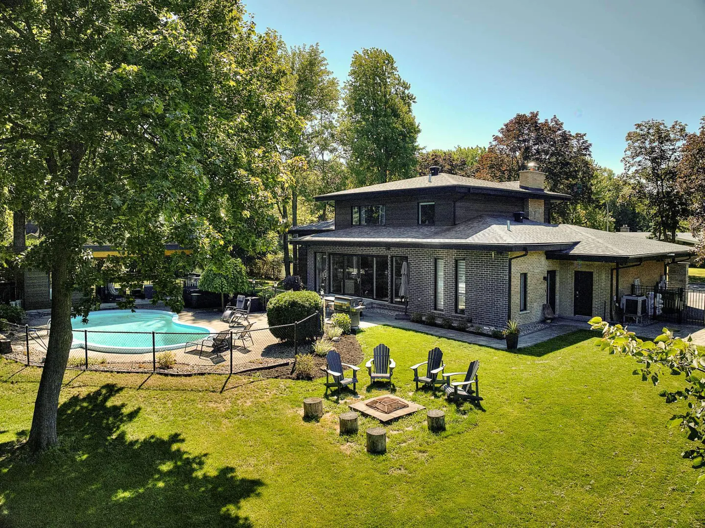 Aerial view of a modern brick house with a pool, fire pit, and green lawn surrounded by trees.