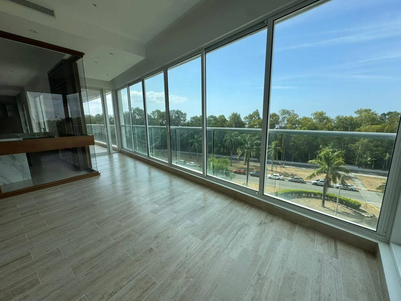 Bright room with beige tile floors and floor-to-ceiling windows. Outside, a balcony overlooks a street with palm trees and greenery.