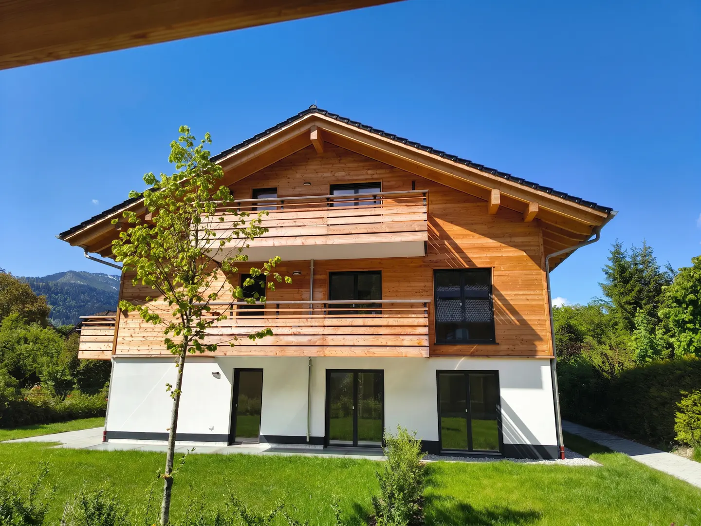 Two-story house with wood siding and a balcony, set against a blue sky and green lawn.