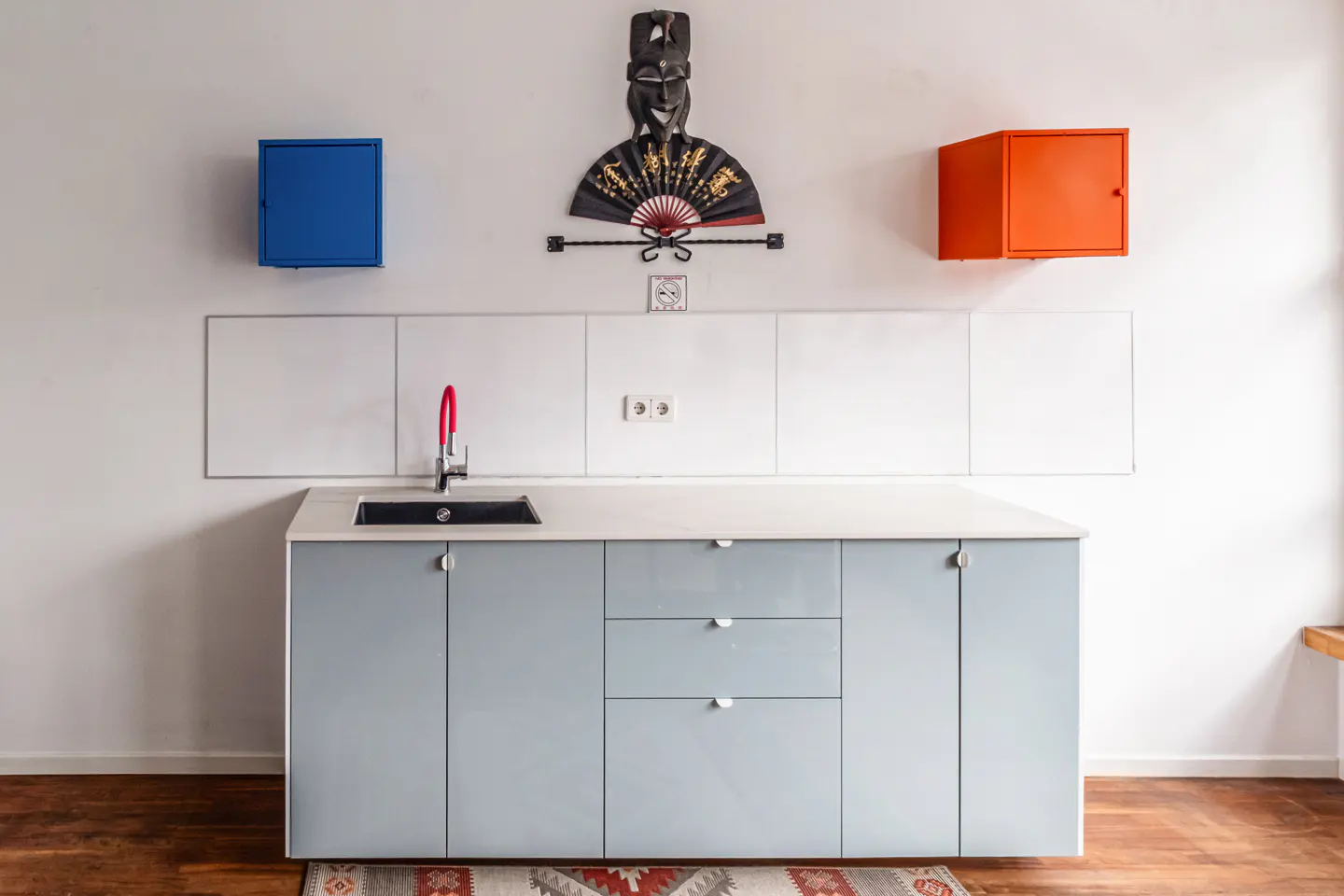 A modern kitchen with light blue cabinets, a red faucet, and colorful wall decor. A patterned rug sits on the wooden floor.