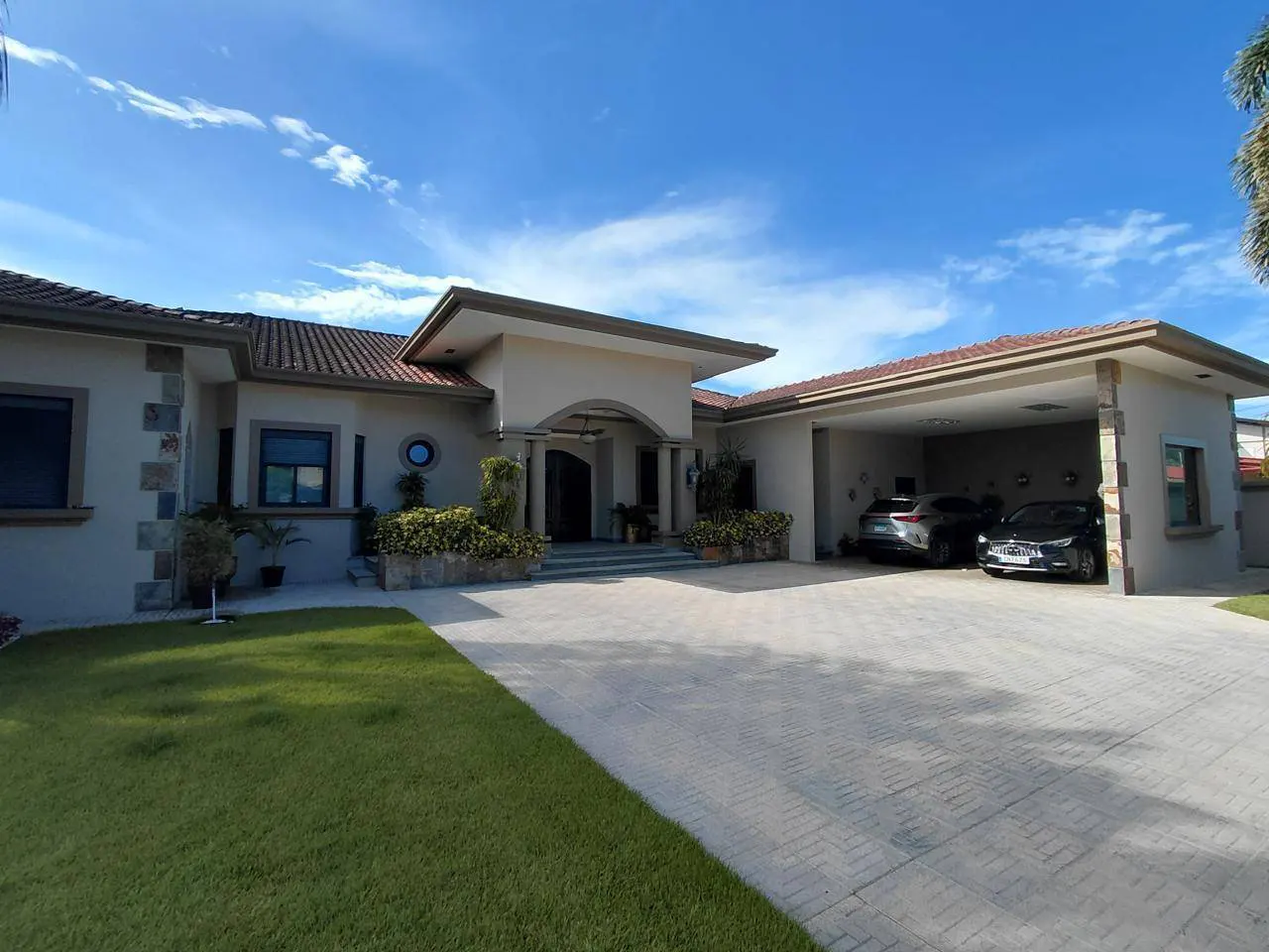 Beige single-story house with a red tile roof, a covered carport with two cars, and a green lawn under a blue sky.