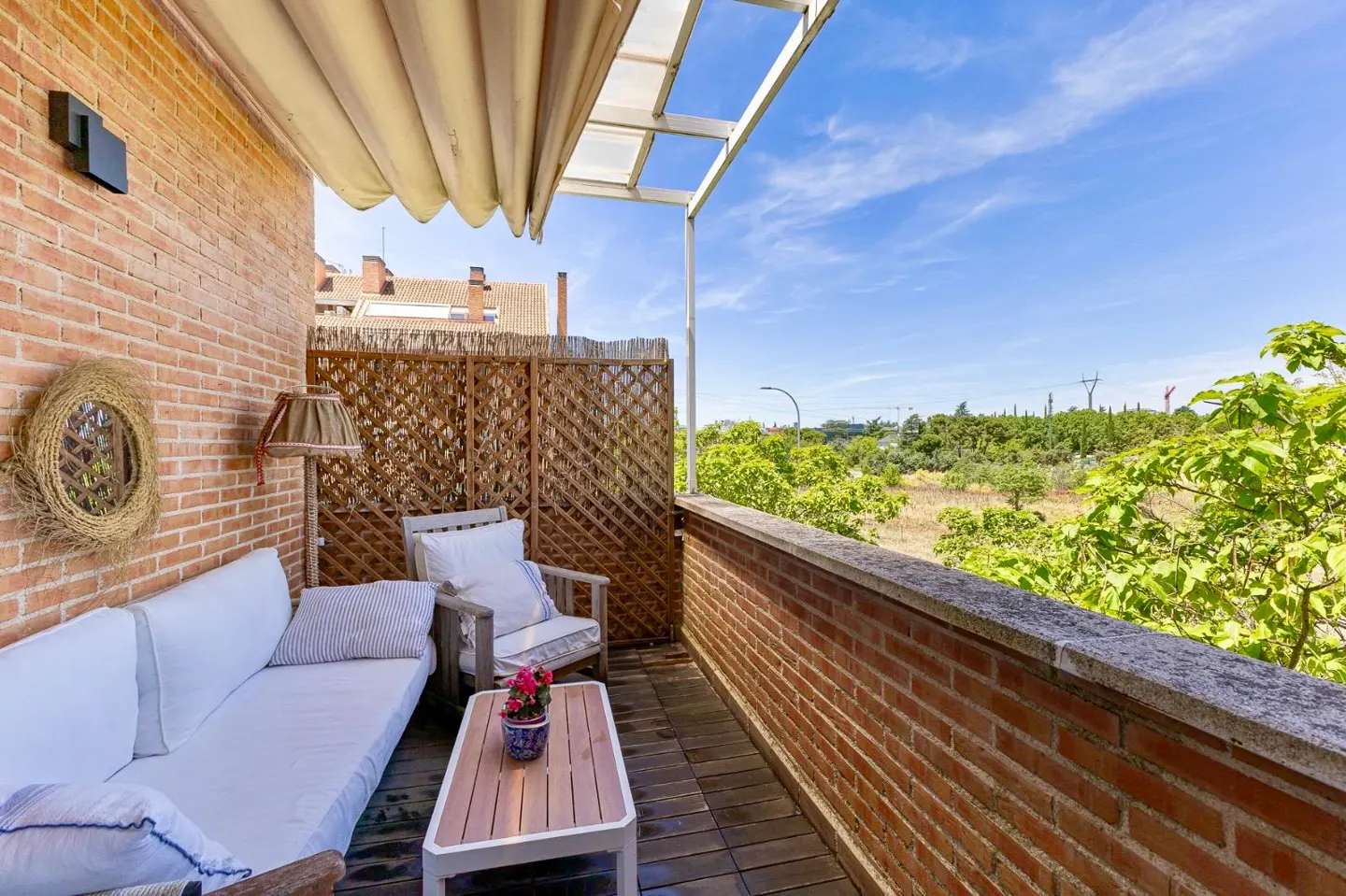 Cozy balcony with a white sofa, wooden coffee table, and lattice privacy screen overlooking a sunny, tree-lined landscape.