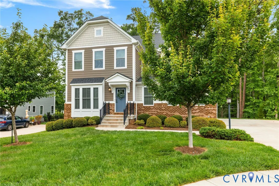 Two-story house with brown siding, brick accents, and a blue front door with a wreath. Green lawn and trees surround the property.