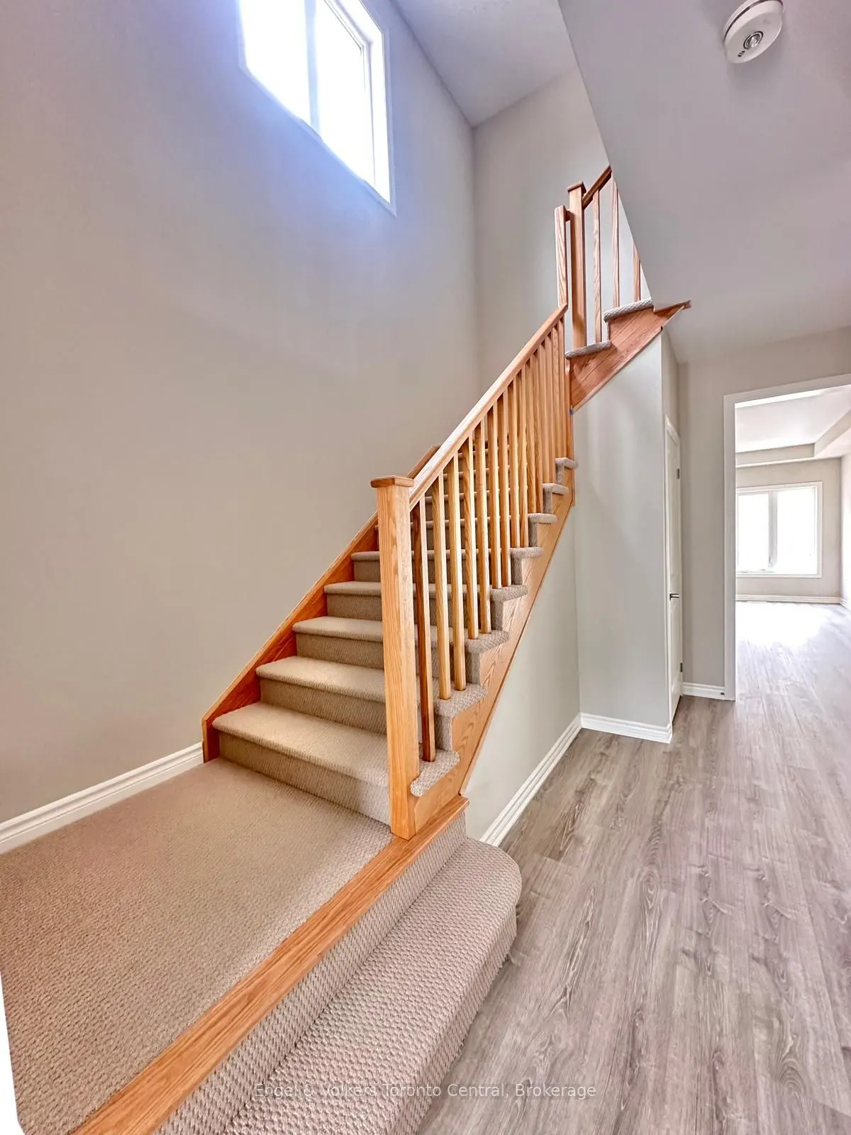 Interior view of a staircase with wood railings and beige carpet, leading to an upper level with a window. Gray wood-look flooring on the ground level.