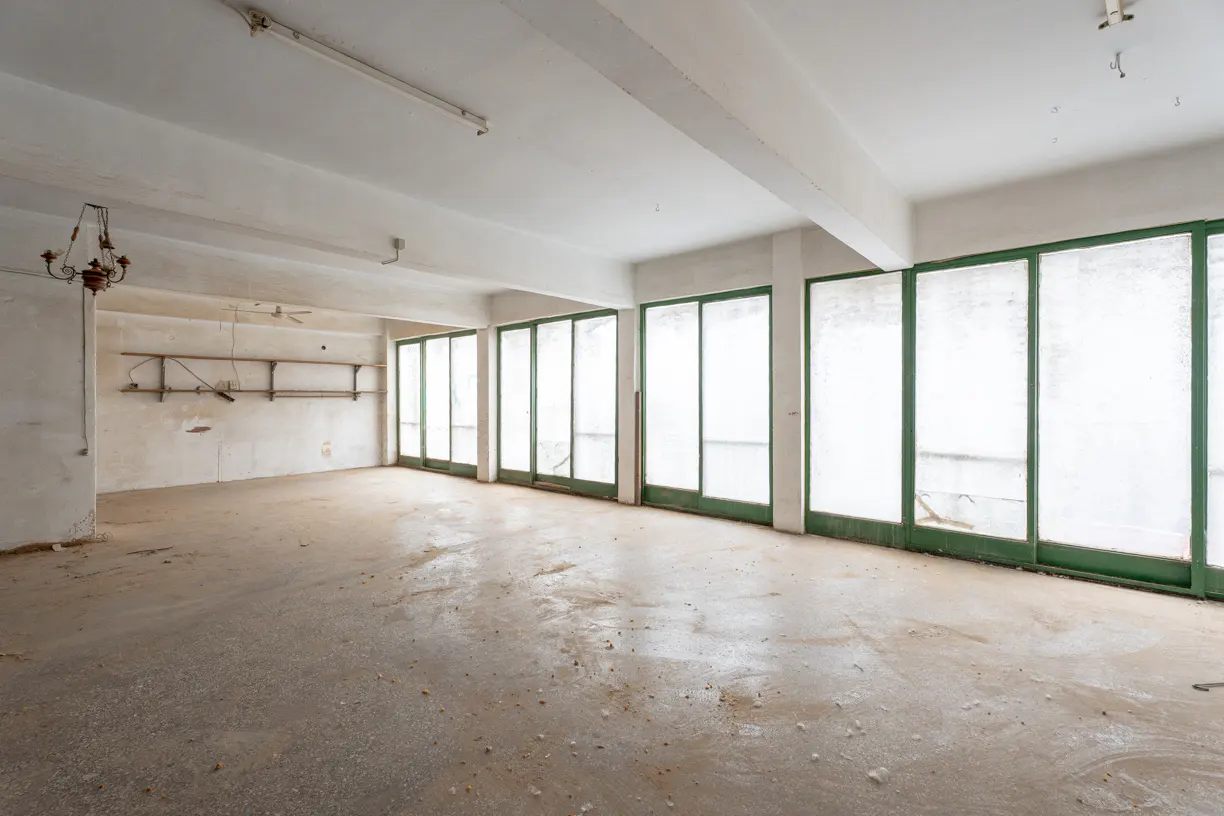 Empty commercial space with a concrete floor, white walls, and green-framed glass doors. A chandelier hangs on the left wall.